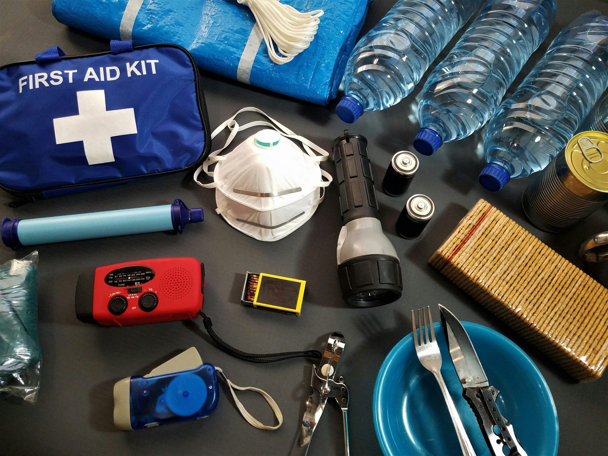 Hurricane emergency preparedness kit with first aid, bottled water, flashlight, hand-crank radio, batteries, dust mask, canned food, and tarp