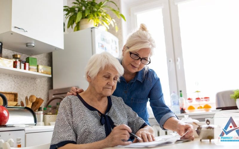 Adult daughter helping her elderly mother review program paperwork at a kitchen table at home